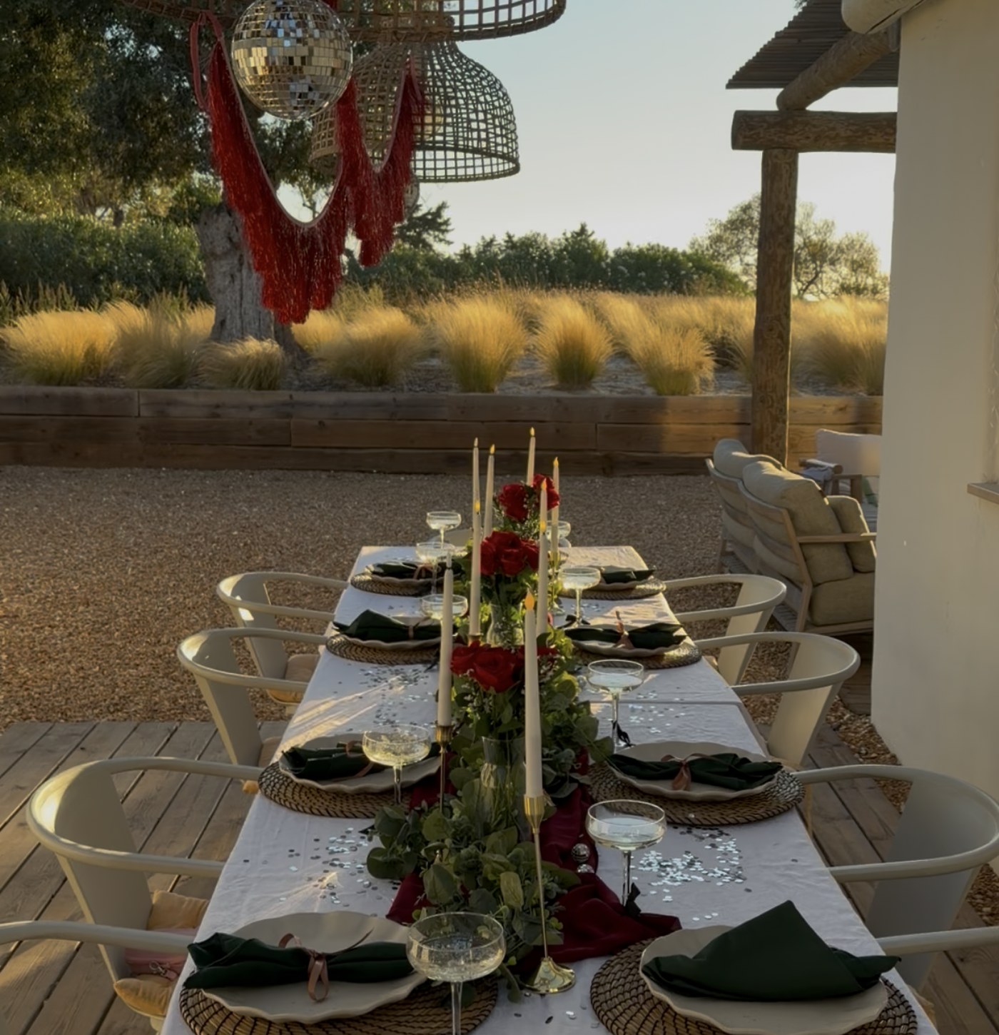 A long table laid for dinner under a pergola at sunset, with candles, red roses and lanterns above
